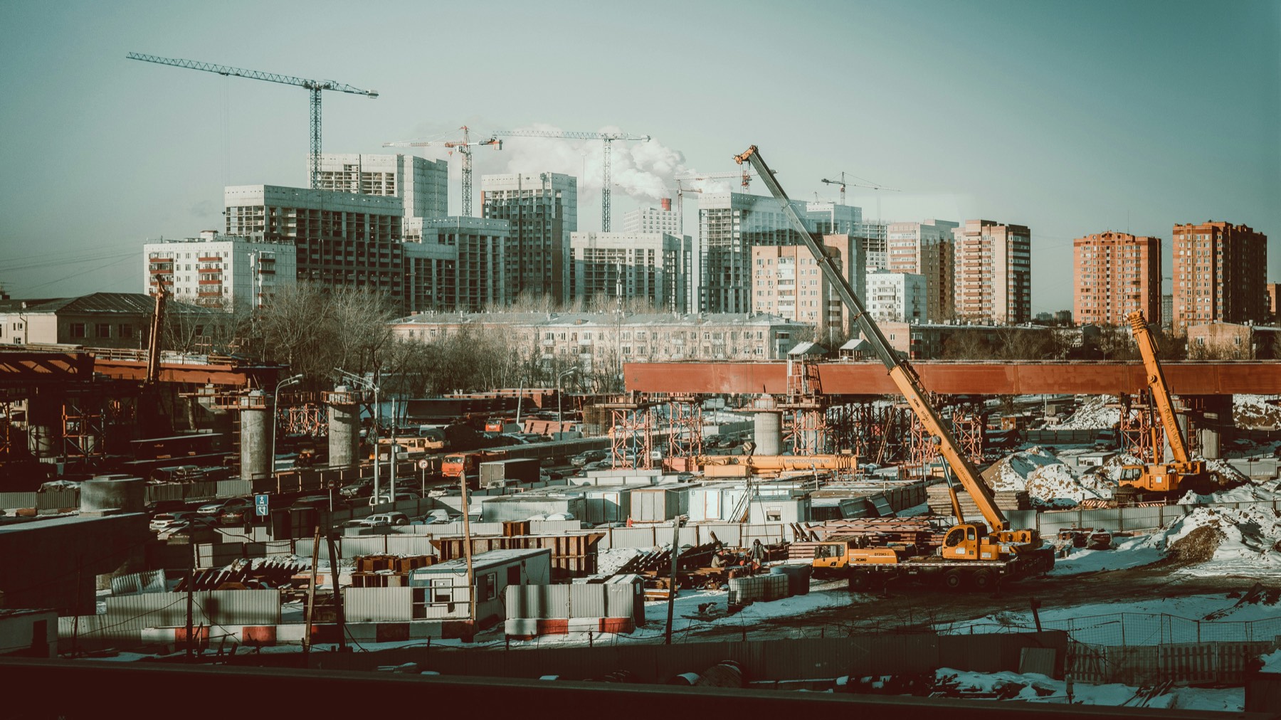 A large active construction site with tower cranes, mobile cranes, and materials, set against a city skyline of buildings under construction